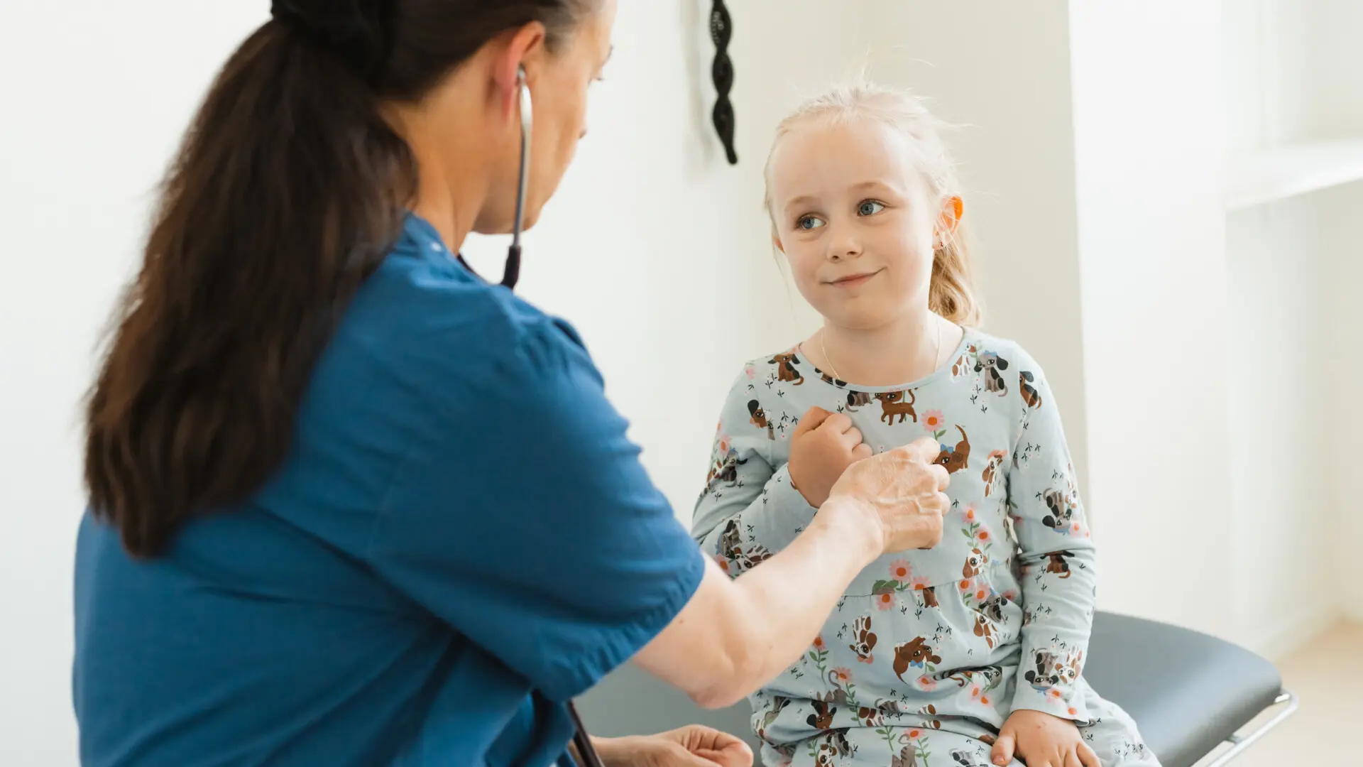 Kvinnlig läkare lyssnar på hjärtat på en liten blond flicka i mönstrad klänning