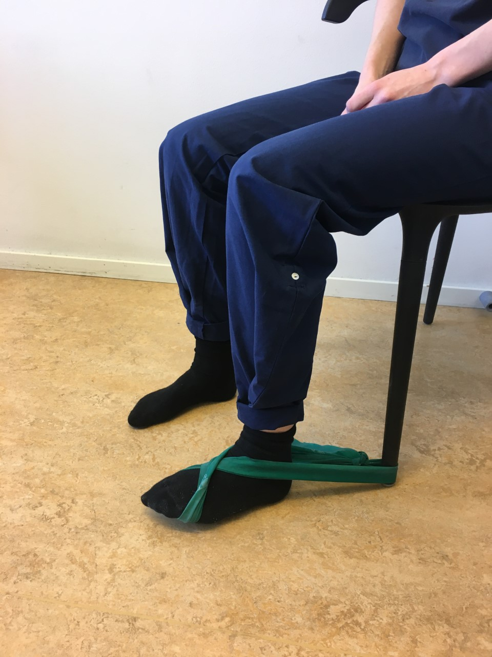 Person sitter p&aring; stol med h&auml;nderna i kn&auml;t. Fr&aring;n stolsbenet och runt foten g&aring;r ett tr&auml;ningsgummiband. Gummibandet g&aring;r i kors &ouml;ver fotryggen och ner under fr&auml;mre delen av foten. 