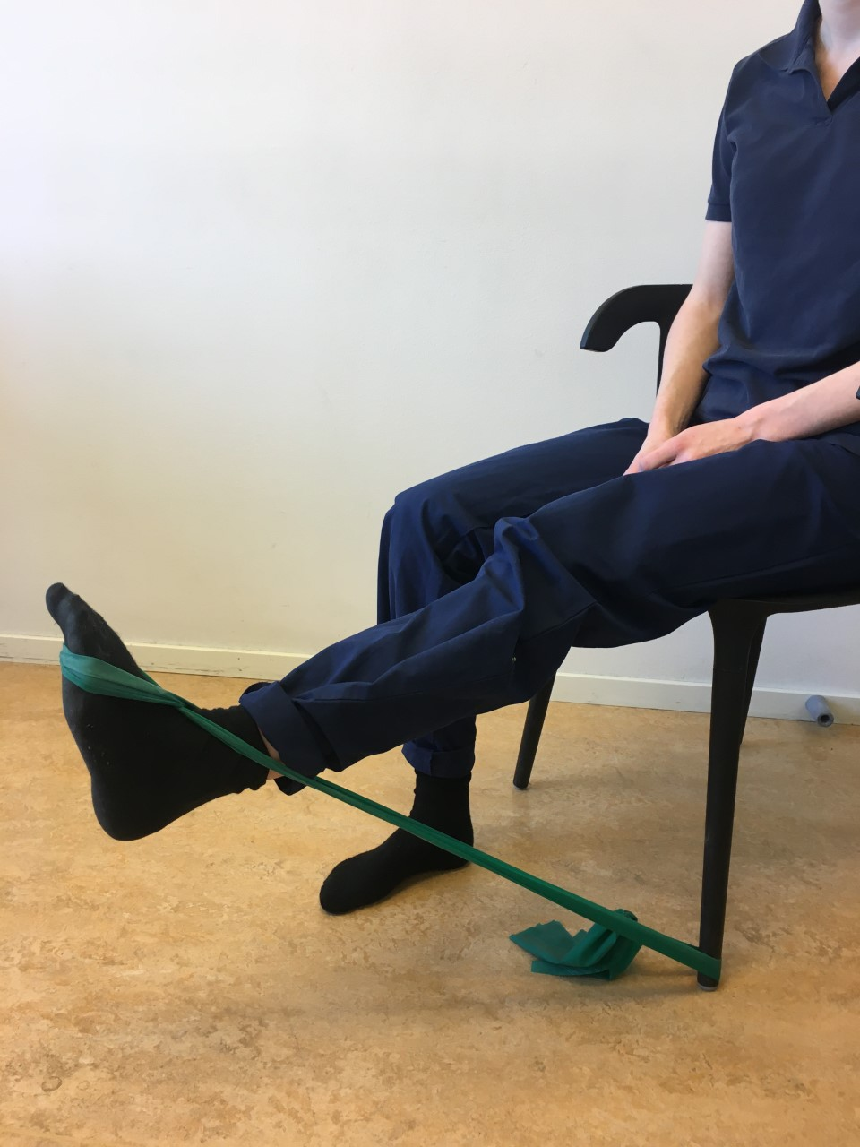 Person sitter p&aring; stol med h&auml;nderna i kn&auml;t. Fr&aring;n stolsbenet och runt foten g&aring;r ett tr&auml;ningsgummiband. Gummibandet g&aring;r i kors &ouml;ver fotryggen och ner under fr&auml;mre delen av foten. Benet str&auml;cks ut&aring;t s&aring; gummibandet t&auml;njs. 