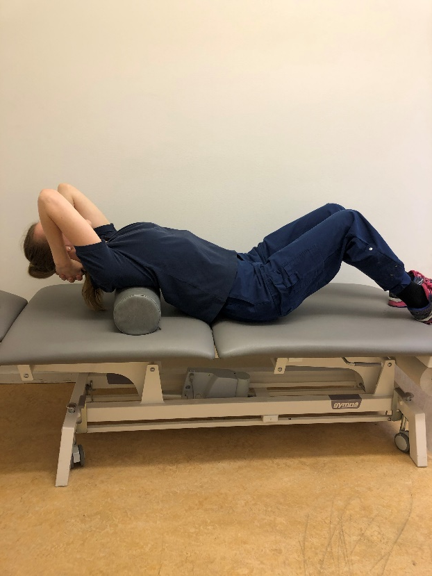 Tjej ligger p&aring; rygg p&aring; en brits med en foamroller uppe vid skulderbladen. Hon har b&ouml;jda ben och h&auml;nderna bakom nacken.