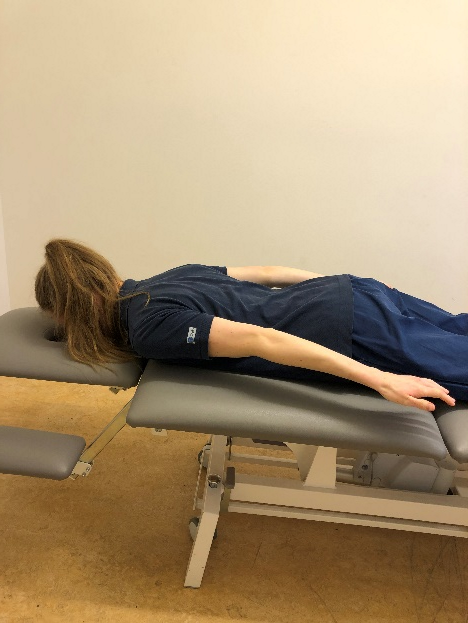 Tjej ligger p&aring; mage p&aring; brits och lyfter raka armar bak&aring;t l&auml;ngs kroppen