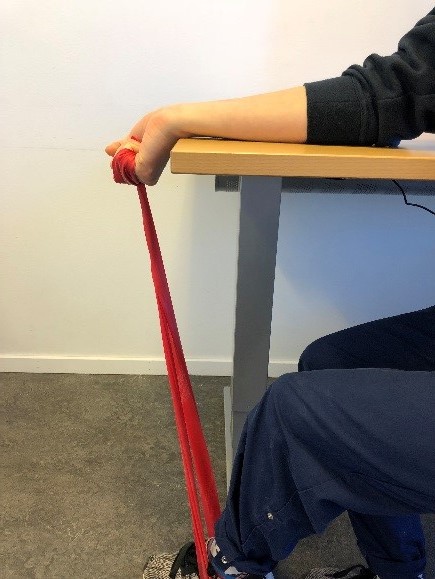 Person sitter vid bord med armen mot ytan, handflatan upp&aring;t och vinklad ner &ouml;ver bordskanten. I den knutna handen ett tr&auml;ningsgummiband som g&aring;r ner till foten som vilar platt mot golvet. 