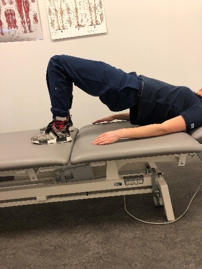 Person gör bäckenlyft på brits. Armarna raka mot underlaget och handflatan mot britsen. Höften lyft.