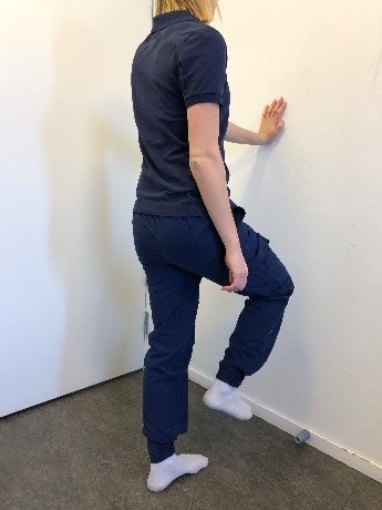 Person står med vänster handflata mot vägg, vänster ben rakt och med hela foten mot golvet. Höger arm hänger avslappnat efter sidan och höger knä är något lyft.