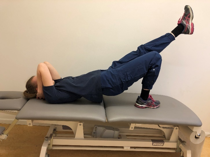 Tjej med ryggen på brits nu med lyft bäcken. Vänster ben är sträckt snett mot taket. Andra med foten uppdragen mot rumpan.