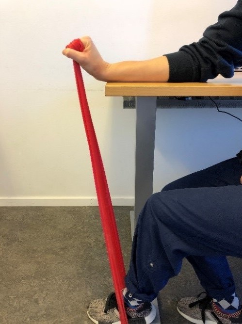 Person sitter vid bord med armen mot ytan, handen knuten, vinklad upp&aring;t. I den knutna handen ett tr&auml;ningsgummiband som g&aring;r ner till foten som vilar platt mot golvet. 