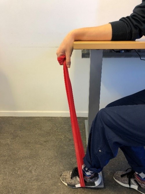 Person sitter vid bord med armen mot ytan, handflatan upp&aring;t och vinklad ner &ouml;ver bordskanten. I den knutna handen ett tr&auml;ningsgummiband som g&aring;r ner till foten som vilar platt mot golvet. 